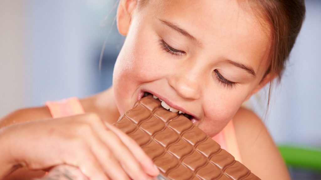 Petite fille souriante croquant une tablette de chocolat au lait, illustrant l’attrait précoce pour le sucre et l’importance de prévenir l’addiction dès l’enfance avec des solutions comme LaserAddict.